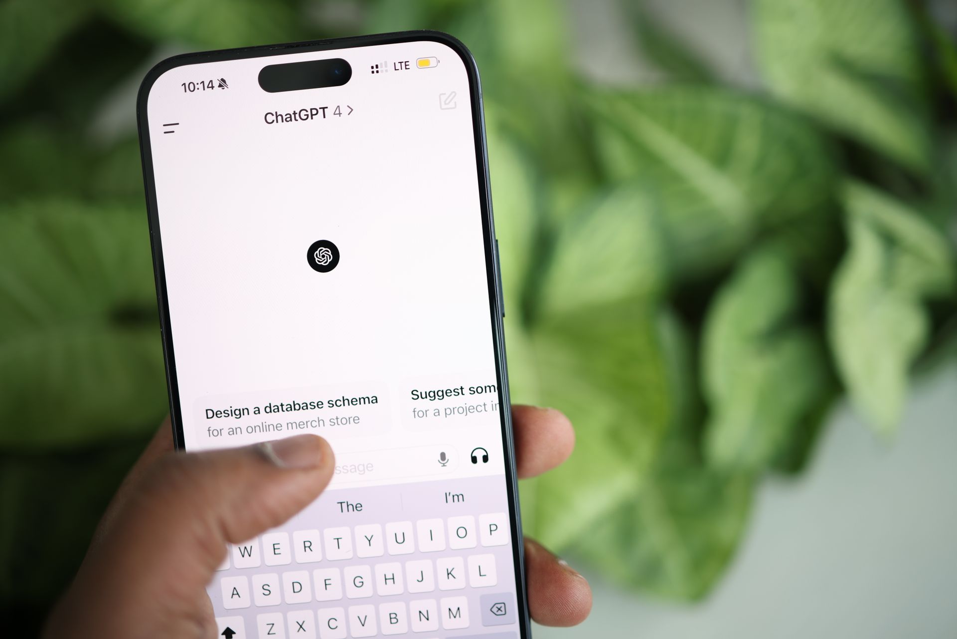 Handy wird in einer Hand gehalten. Der Bildschirm zeigt die Startseite von ChatGPT. Im Hintergrund sieht man einen Schreibtisch und Pflanzen.